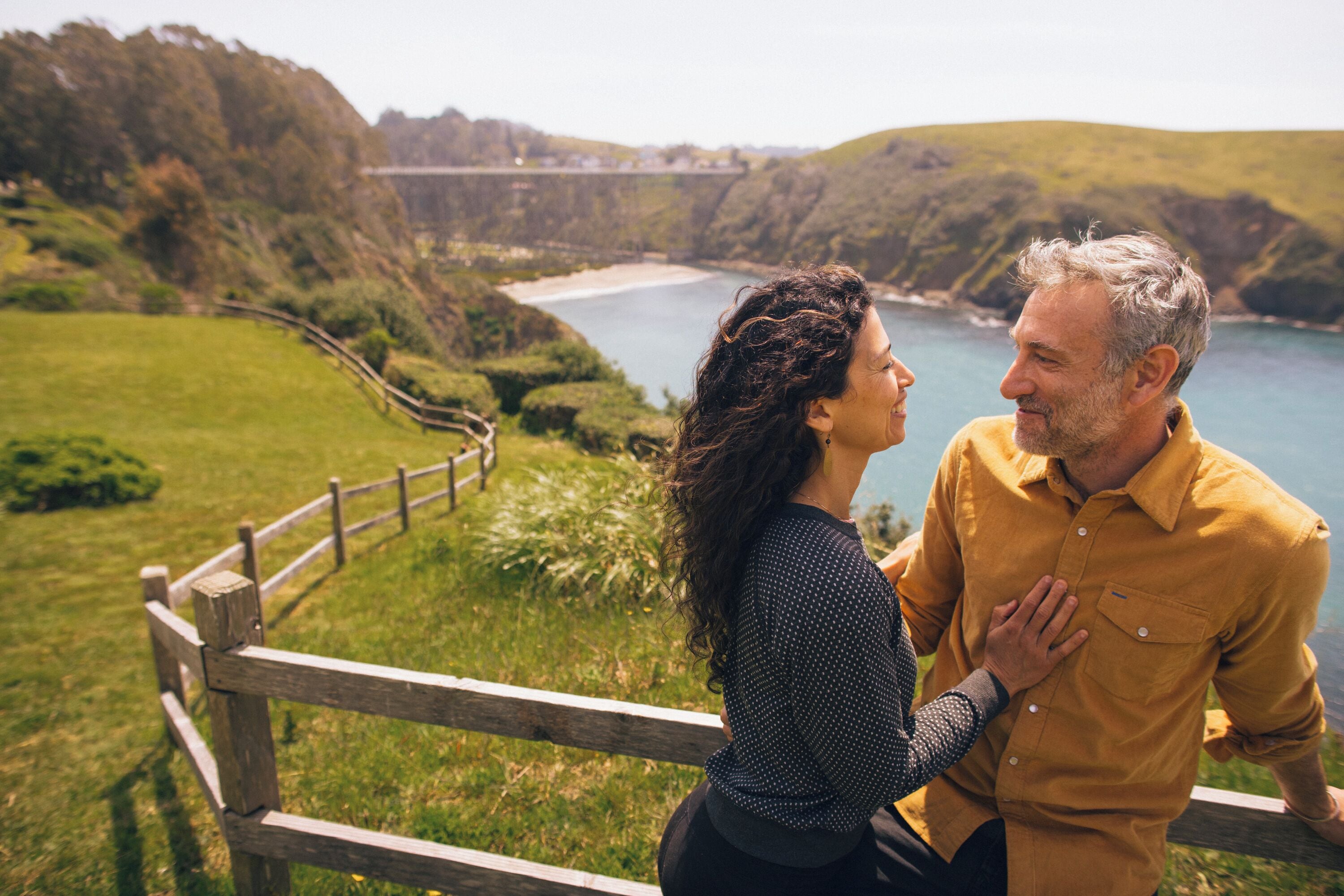 A couple sitting on a wooden bench overlooking a coastal landscape with hills and a bridge in the distance.