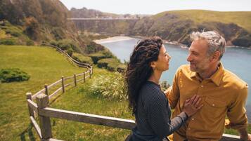 A couple sitting on a wooden bench overlooking a coastal landscape with hills and a bridge in the distance.
