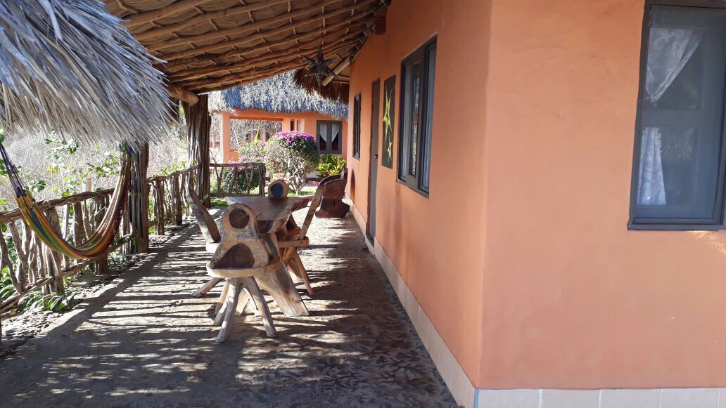 A covered outdoor area with a wooden table and chairs, a thatched roof, and a building with windows.