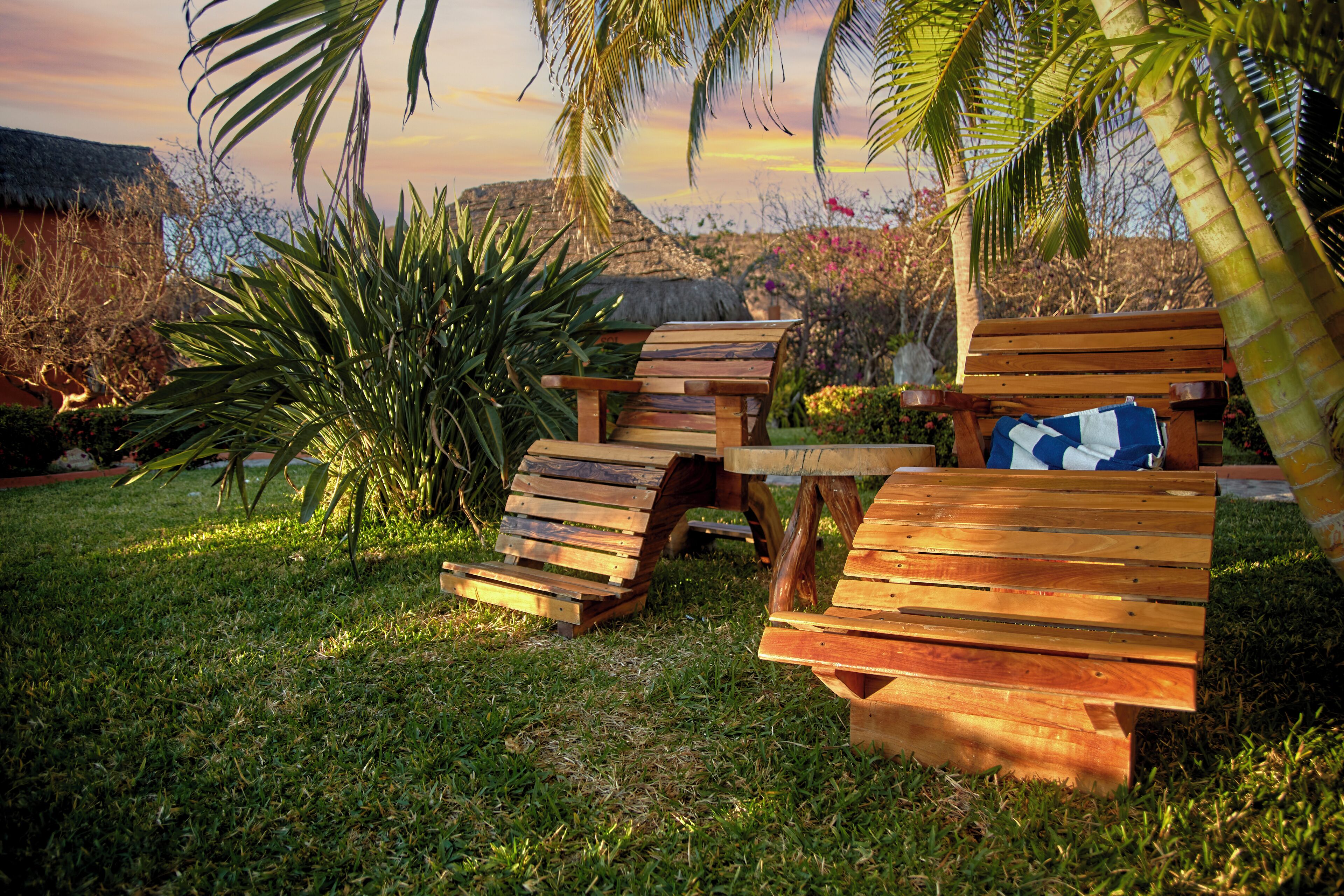Un conjunto de banco y mesa de madera en un área cubierta de césped, con palmeras y una estructura de techo de paja al fondo.