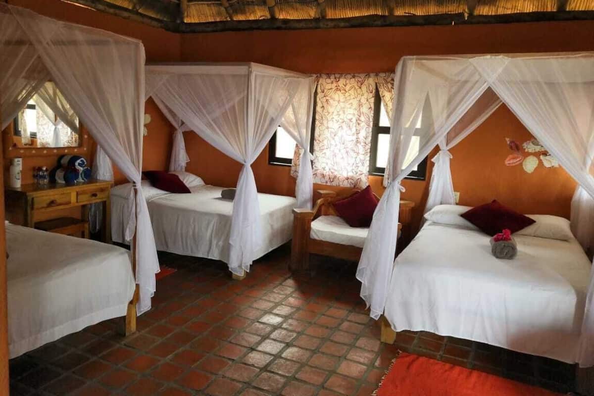 Une chambre avec quatre lits, chacun surmonté d’un dais. On y trouve une table de chevet en bois avec un miroir, ainsi qu’une petite table avec une lampe. La chambre a un sol carrelé et un toit de chaume.