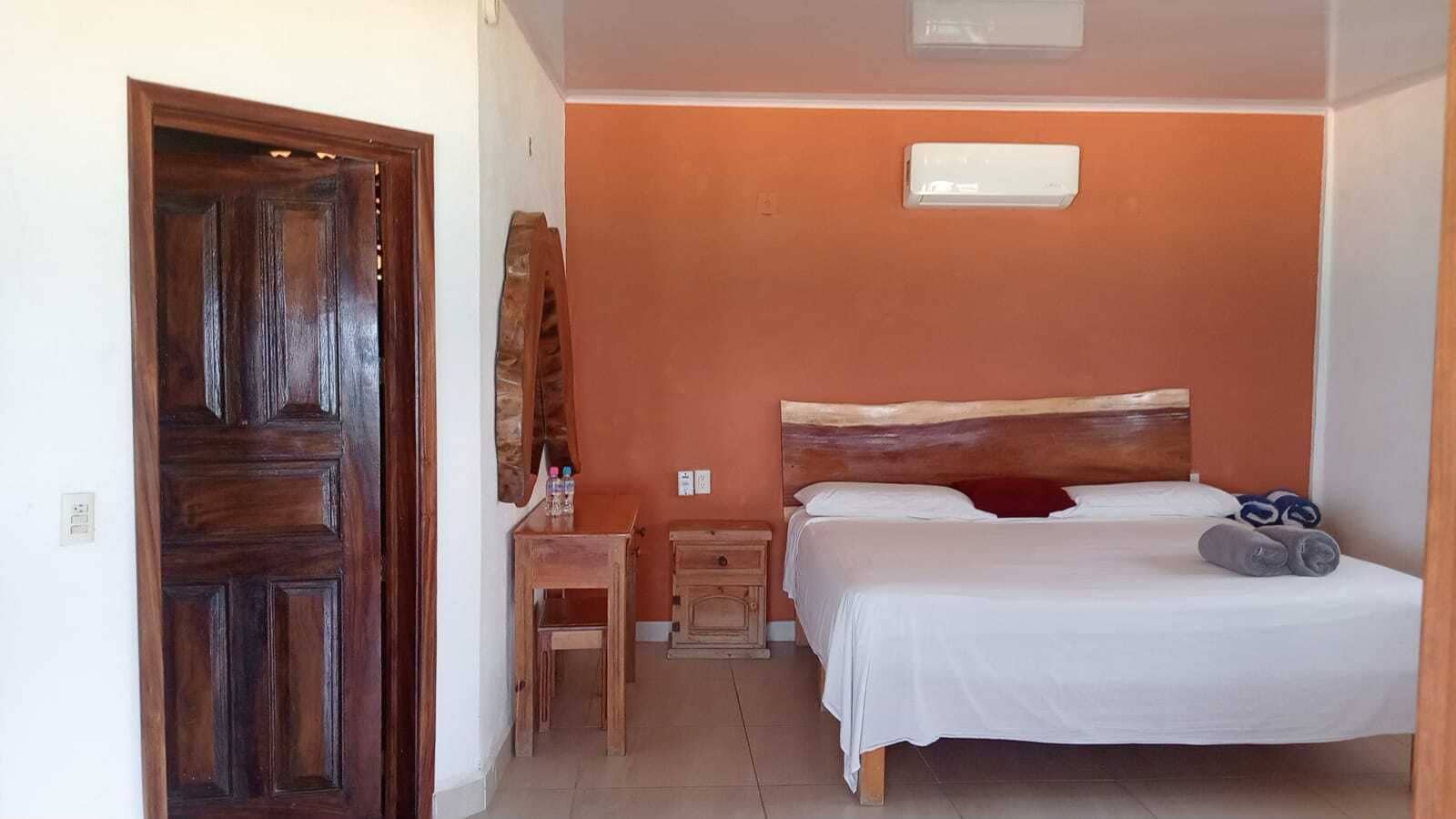 Une chambre avec une porte en bois, une petite table en bois sur laquelle sont posés des articles de toilette, un lit avec du linge de lit blanc et une table de chevet.