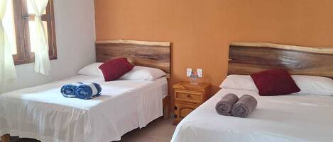 Dos camas individuales con cabeceros de madera, ropa de cama blanca y almohadas rojas. Hay una mesita de noche de madera con una botella y un vaso. Un equipo de aire acondicionado está montado en la pared.