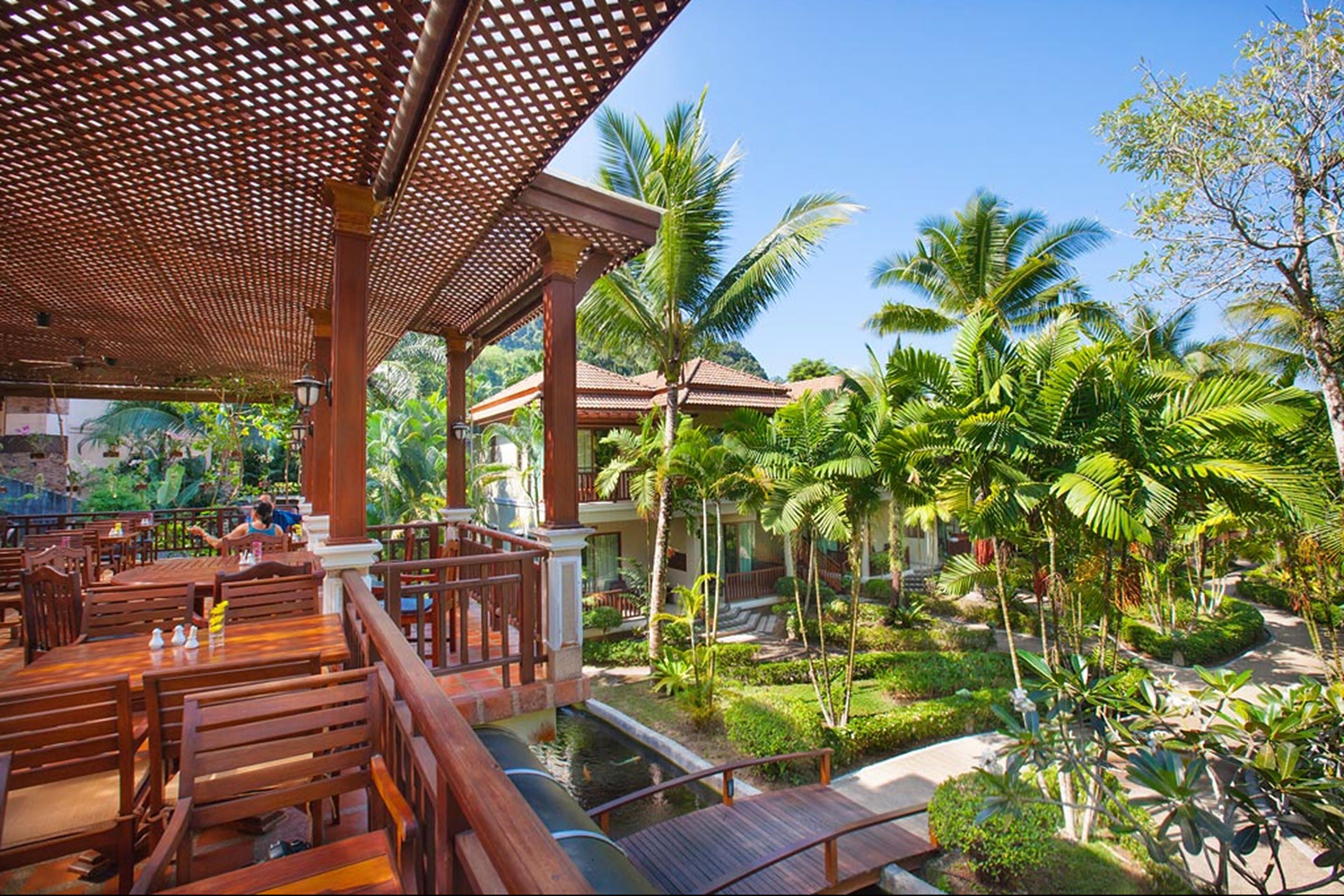 A restaurant with wooden tables and chairs, a covered outdoor seating area, and a view of palm trees and a water feature.