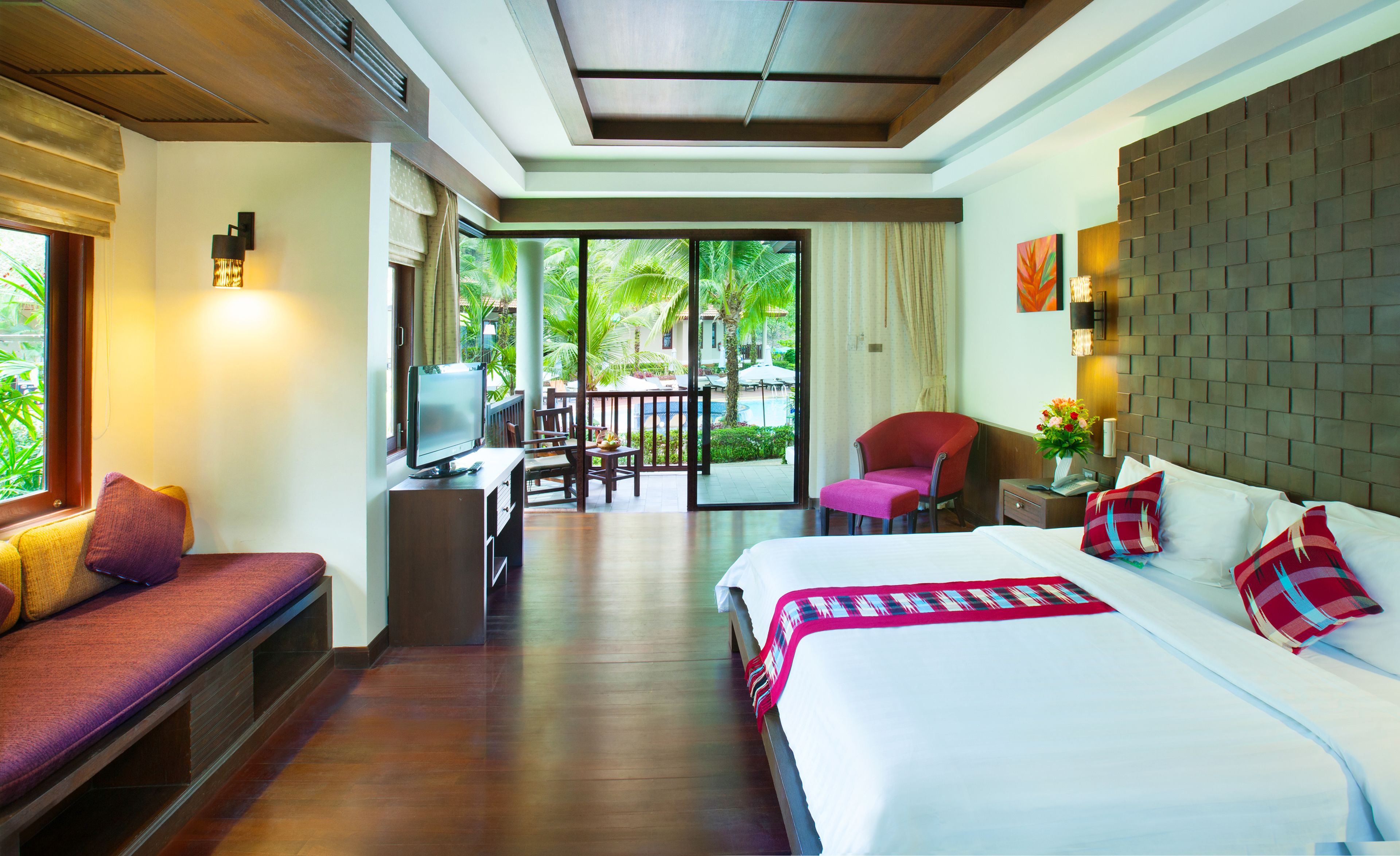 A hotel room with a bed, a desk with a computer, a chair, a sofa, and a view of a pool and palm trees.