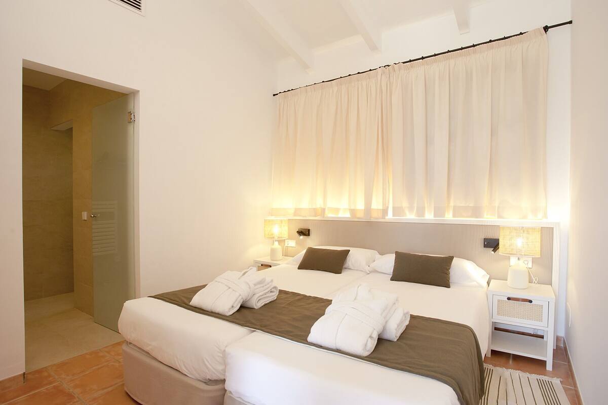 A hotel room with a double bed, two bedside tables with lamps, a window with sheer curtains, and a door leading to a bathroom.