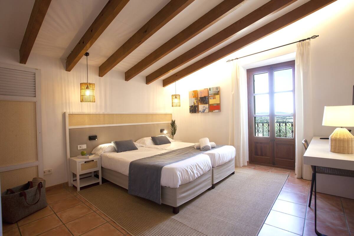 A bedroom with a large bed, a bedside table, a lamp, a door with a view, and a small desk with a lamp.