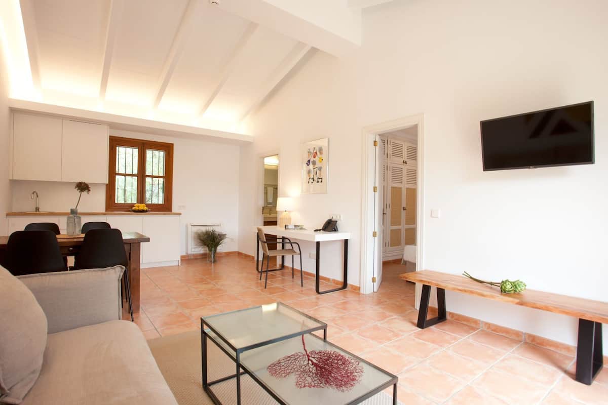 A spacious living area with a glass coffee table, a sofa, a dining table with chairs, a kitchen with a window, and a TV mounted on the wall.