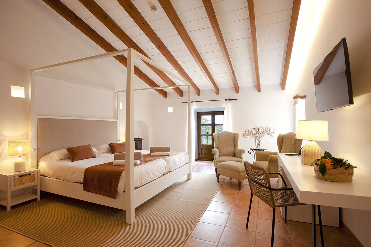 A bedroom with a four-poster bed, a desk with a chair, a TV, and a window with curtains.