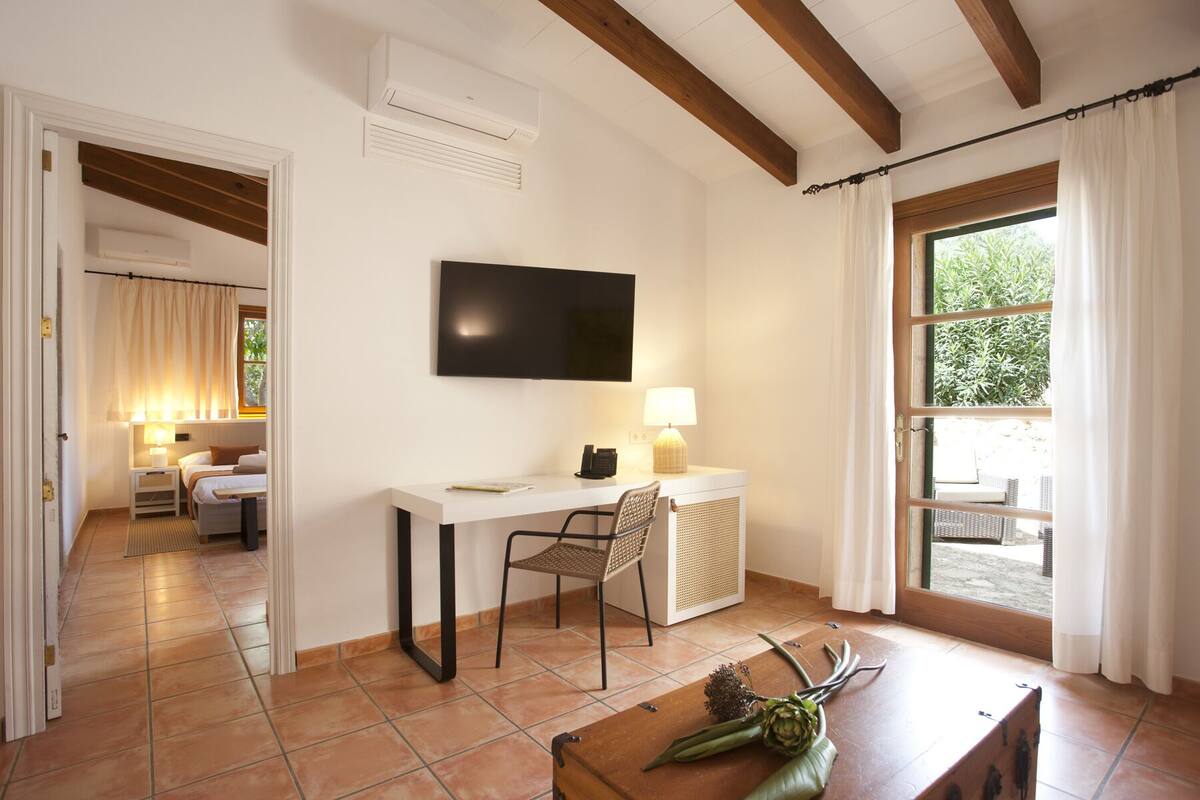 A hotel room with a desk, chair, and a TV mounted on the wall. There is a window with curtains, a door leading to another room, and a view of the outdoors.