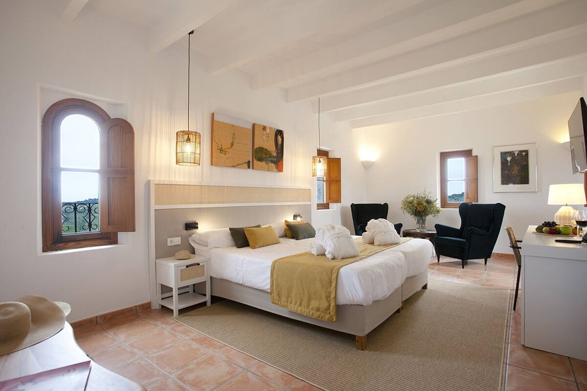 A spacious bedroom with a large bed, bedside tables, a desk, and a chair. There are artwork on the walls and a window with a view.