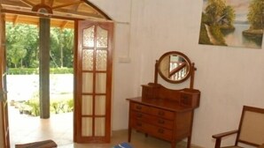 A room with a wooden dresser, a chair, a mirror, and a view of a garden through an open door. - The Bungalow at Pantiya Estate (Matugama)