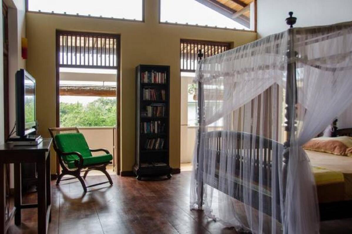 A room with a bed, a green chair, a bookshelf, and a window with a view of greenery.