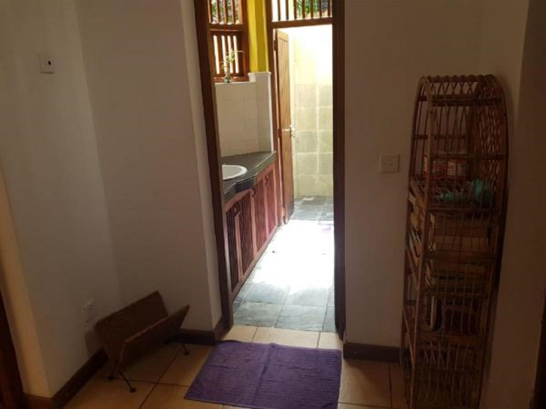 A narrow hallway with a purple mat, a wooden bench, a kitchen area with a sink, and a wicker basket.