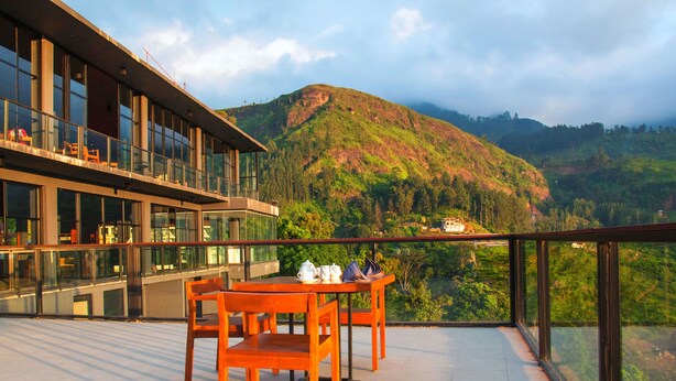 A modern building with a balcony, a table with chairs, and a scenic view of a hill. - Oak Ray Tea Bush (Kotmale)