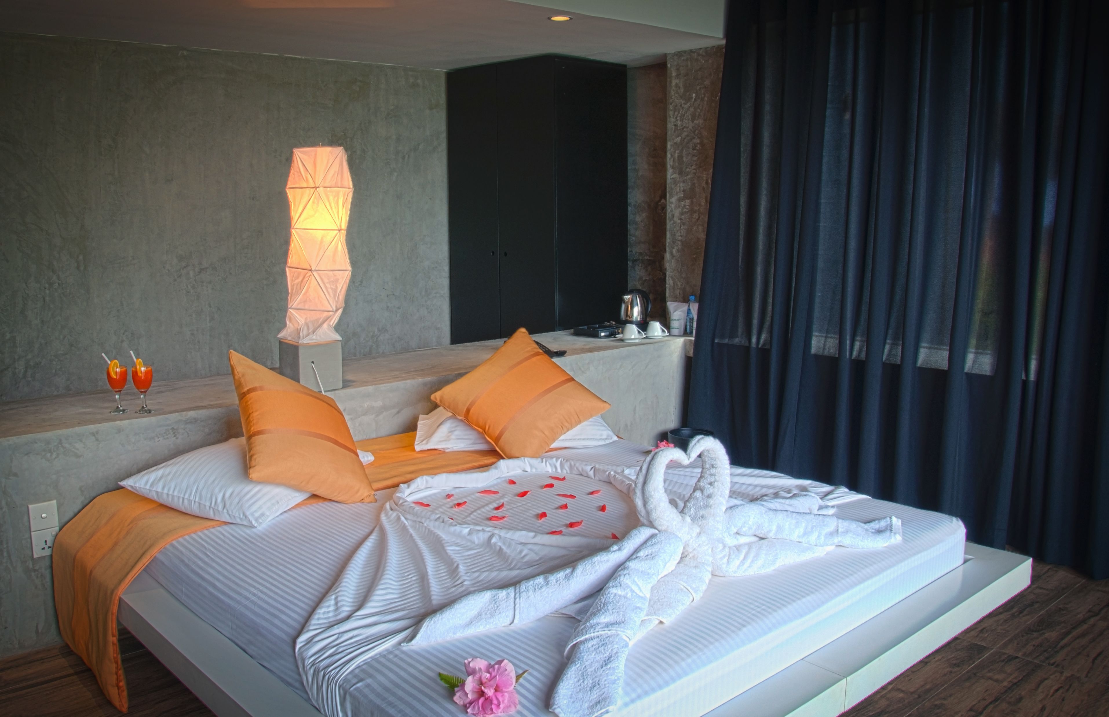 A modern hotel room with a large bed, orange pillows, a decorative wall lamp, and a view of the city through dark curtains.