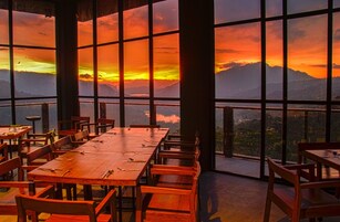 Restaurante com grandes janelas proporcionando vista para montanhas e um pôr do sol.