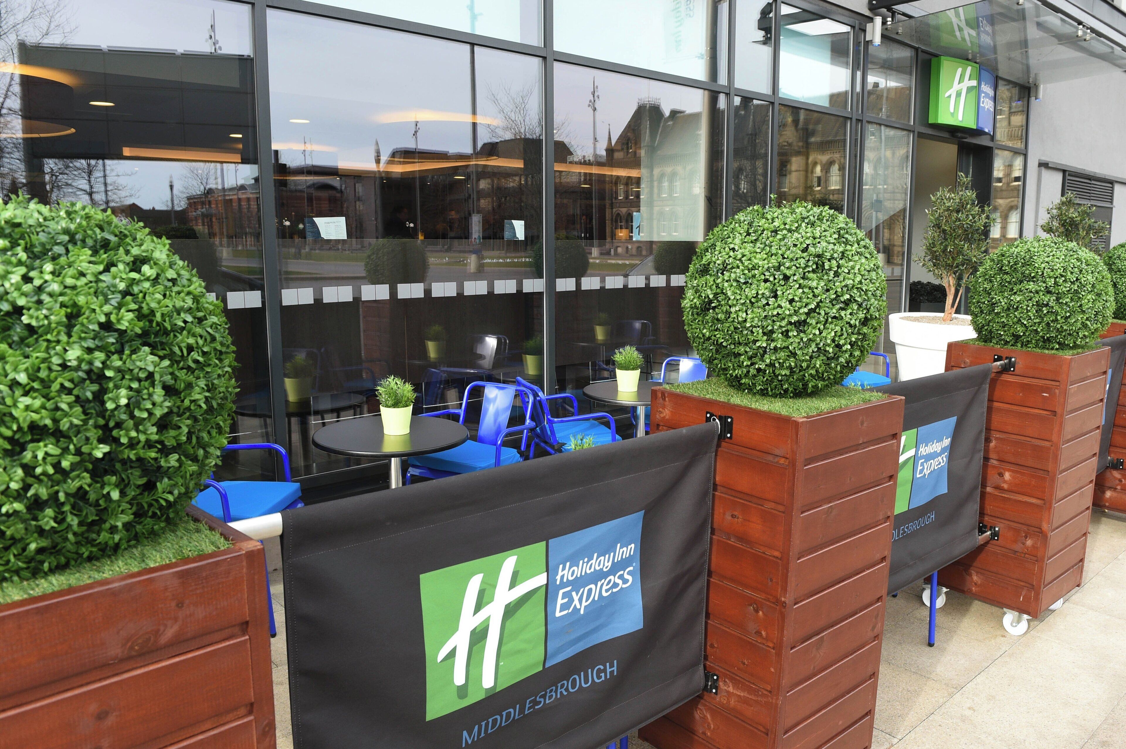 Sitzbereich im Freien mit einem Schild von Holiday Inn Express, Blumentöpfen und einem Ziegelpflanzkübel.