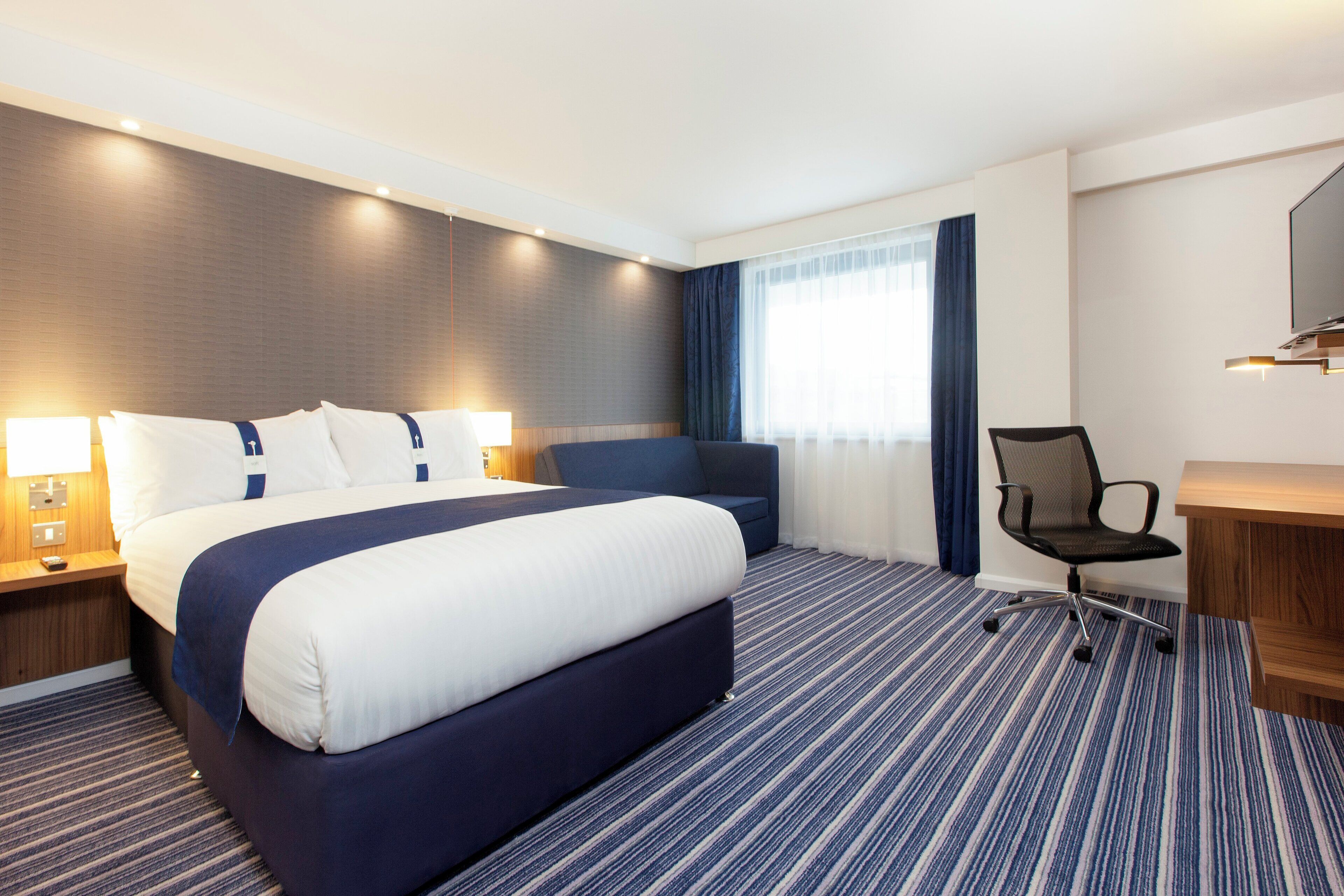 A hotel room with a large bed, a desk, a chair, and a window with curtains.