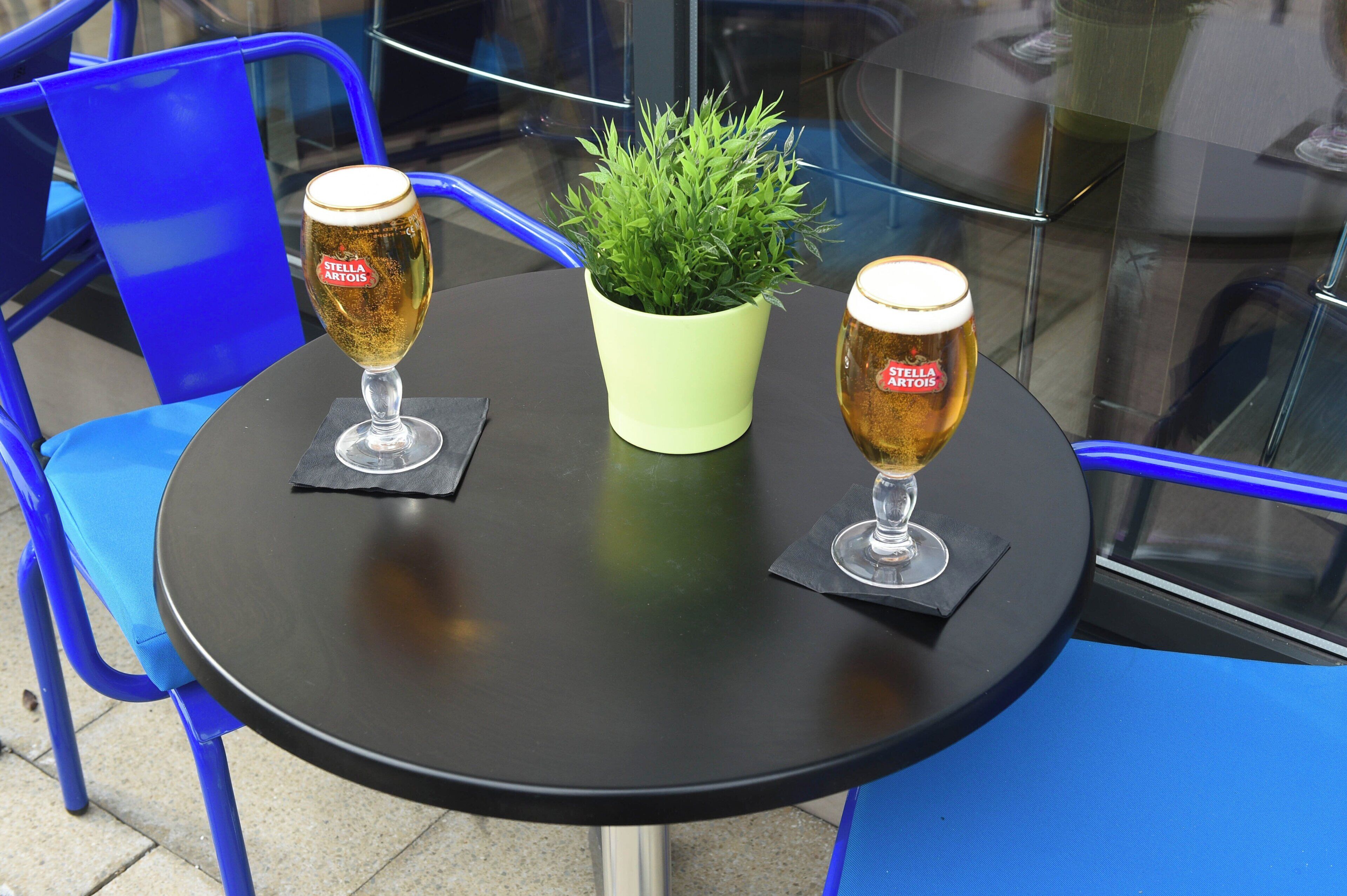 Zwei Biergläser auf einem runden Tisch, dazwischen ein Blumentopf, alles auf einem blauen Stuhl platziert.