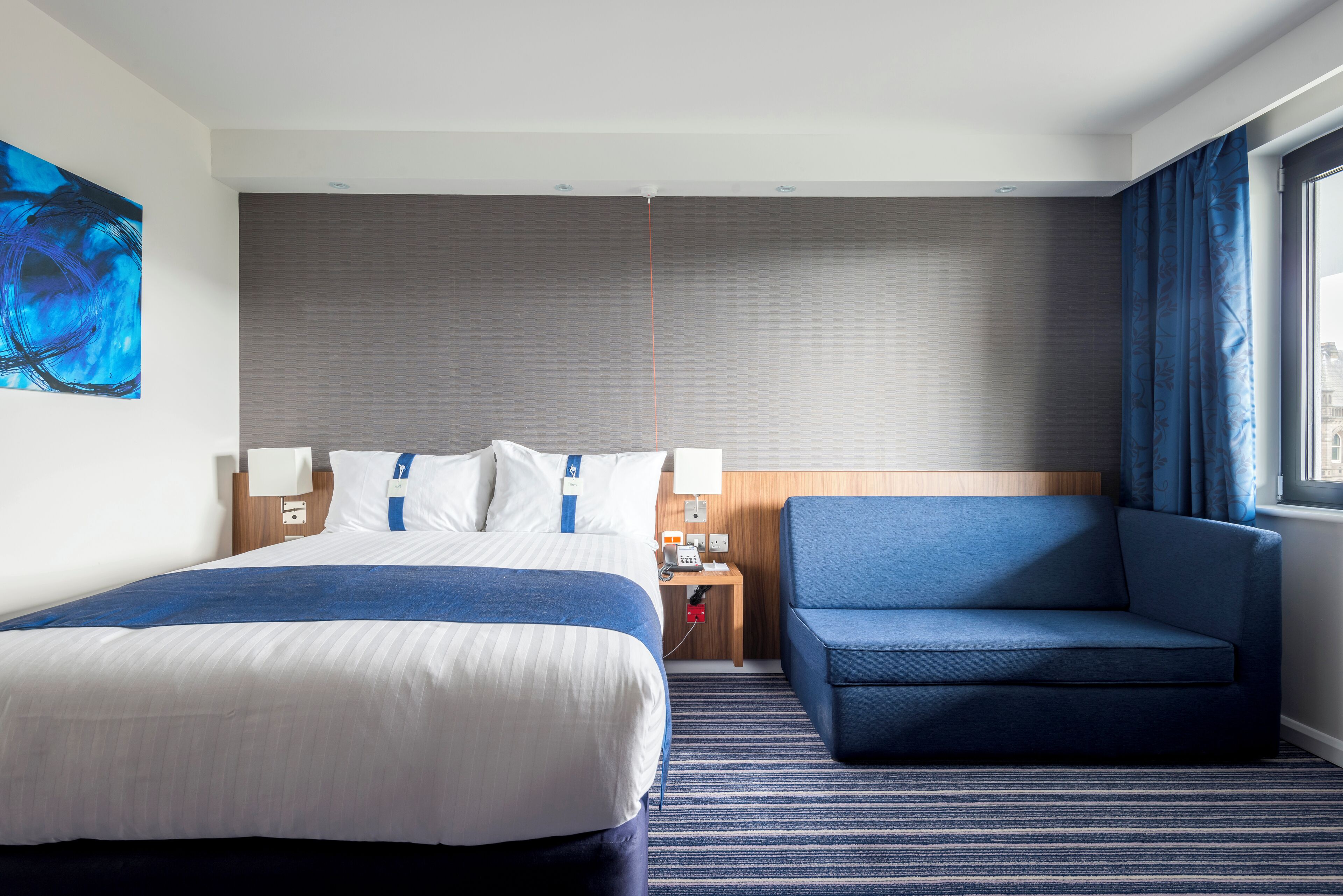 A hotel room with a large bed, a blue sofa, a nightstand with a lamp, and a painting on the wall.
