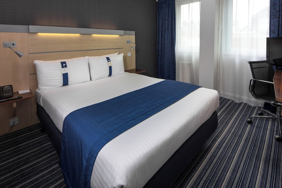 Una camera d'albergo con un letto grande, una scrivania con una sedia, una lampada e una finestra con tende.