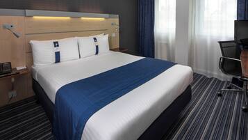Habitación de hotel con una cama grande, un escritorio con silla, una lámpara y una ventana con cortinas.