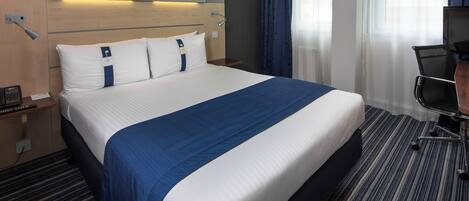Habitación de hotel con una cama grande, un escritorio con silla, una lámpara y una ventana con cortinas.