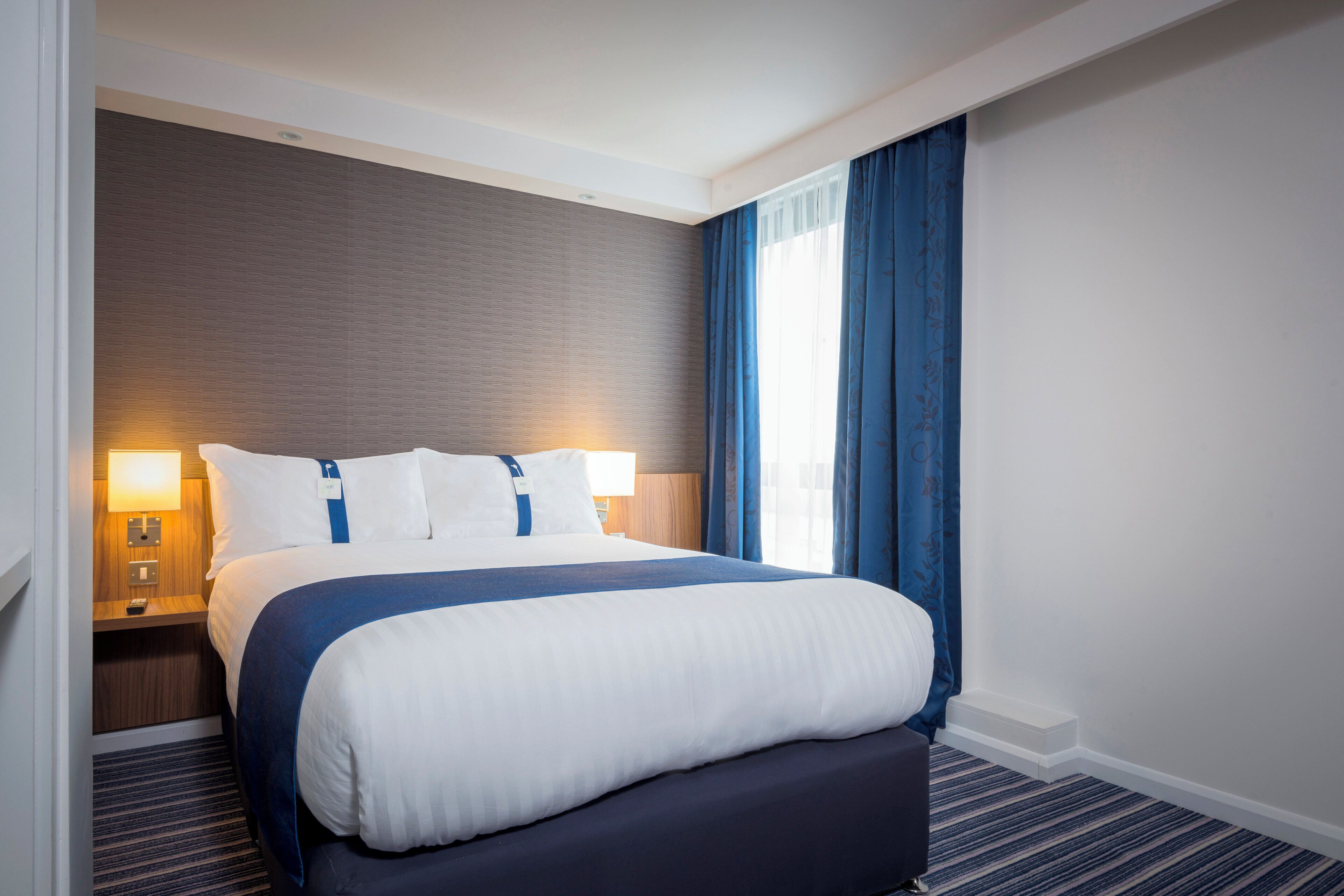 A hotel room with a bed, a nightstand, a lamp, and a window with curtains.