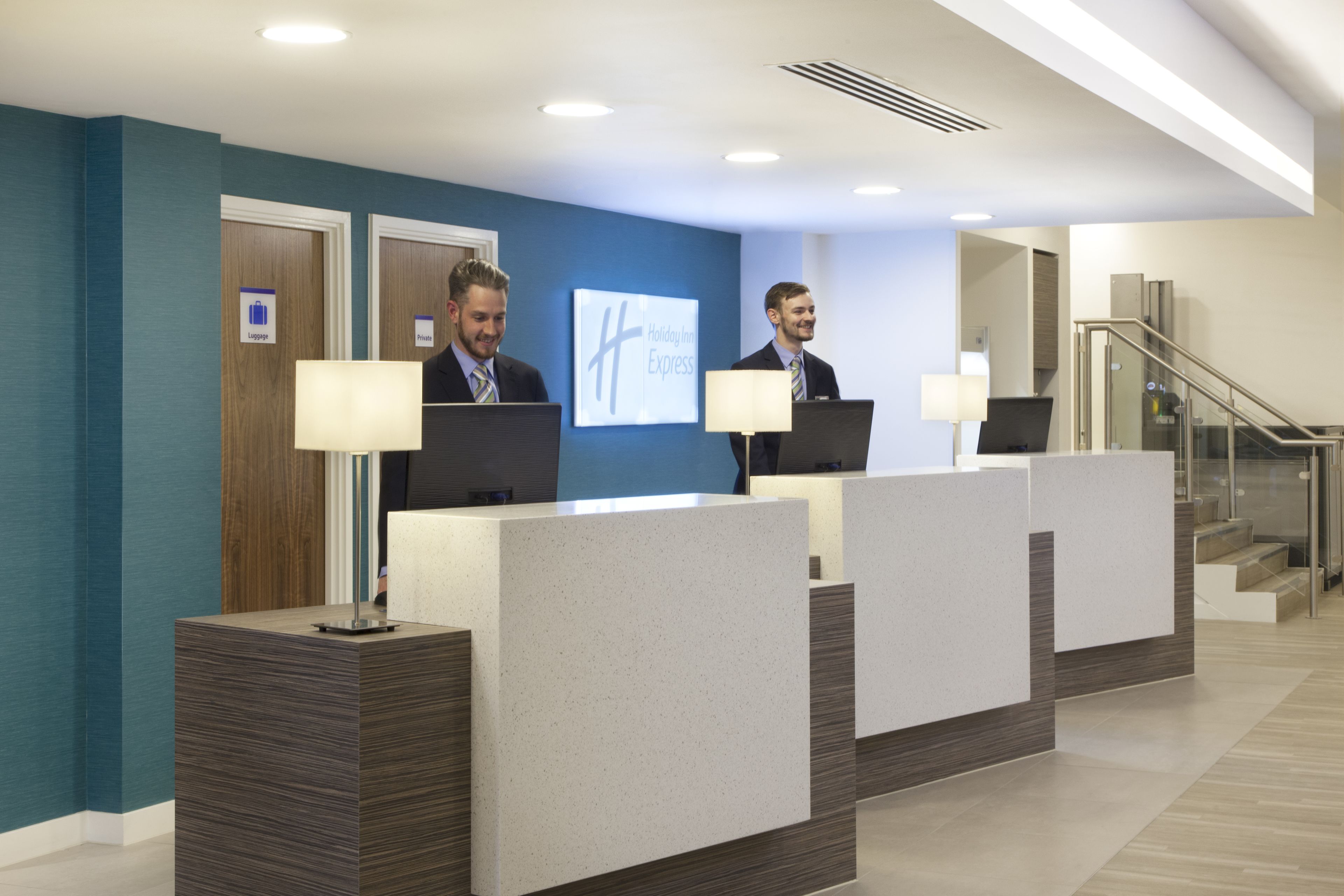 Zona de recepción de hotel con pared azul, un cartel de Holiday Inn Express y una escalera a la derecha.