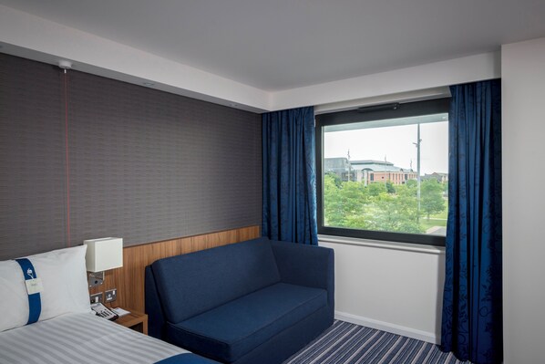 A hotel room with a bed, a sofa, a window with curtains, a lamp, and a telephone.
