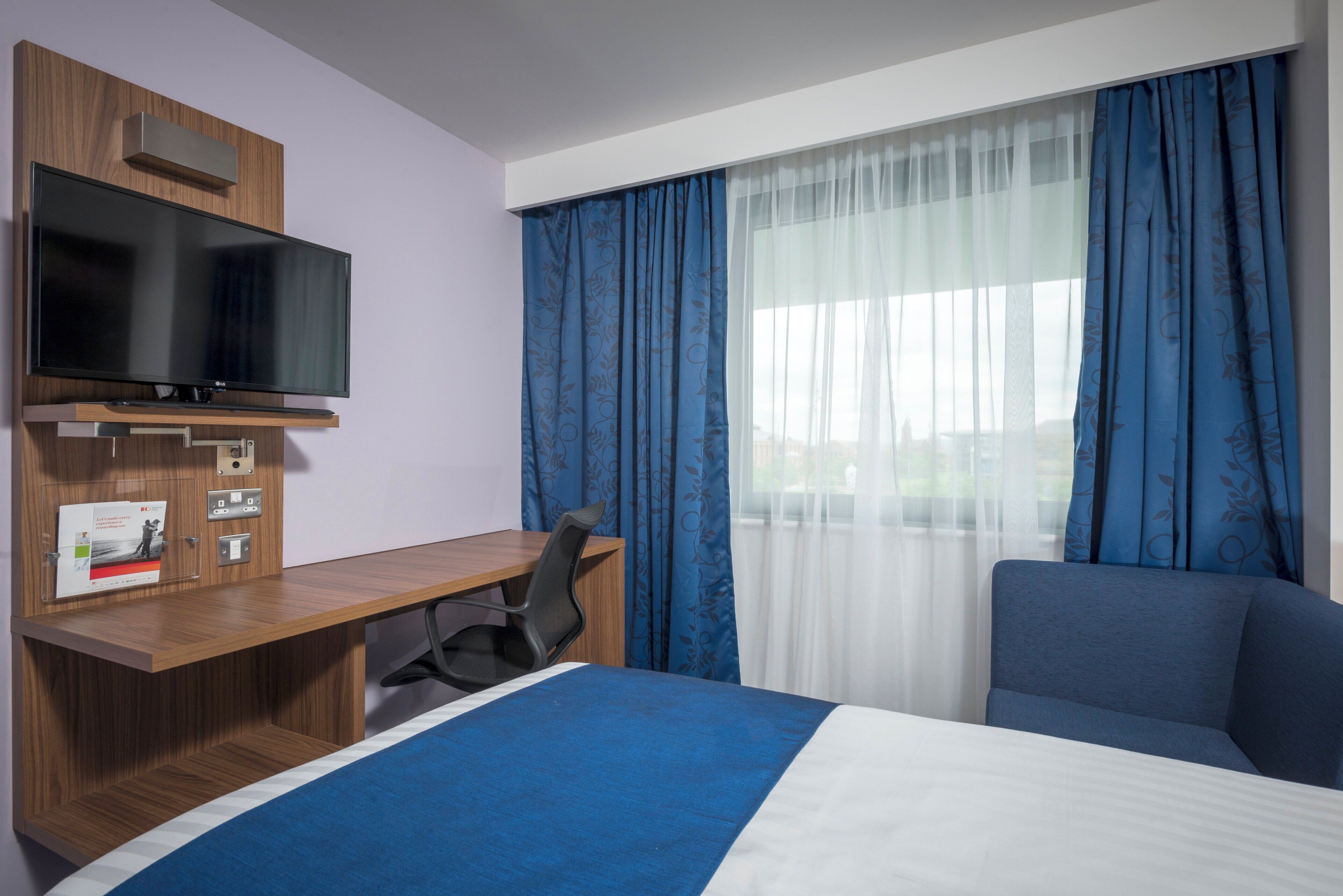 A hotel room with a bed, a desk with a chair, a television, and a window with curtains.