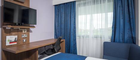 A hotel room with a bed, a desk with a chair, a television, and a window with curtains.