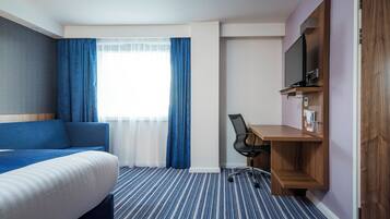 A hotel room with a bed, a desk, a chair, a TV, and a window with curtains.