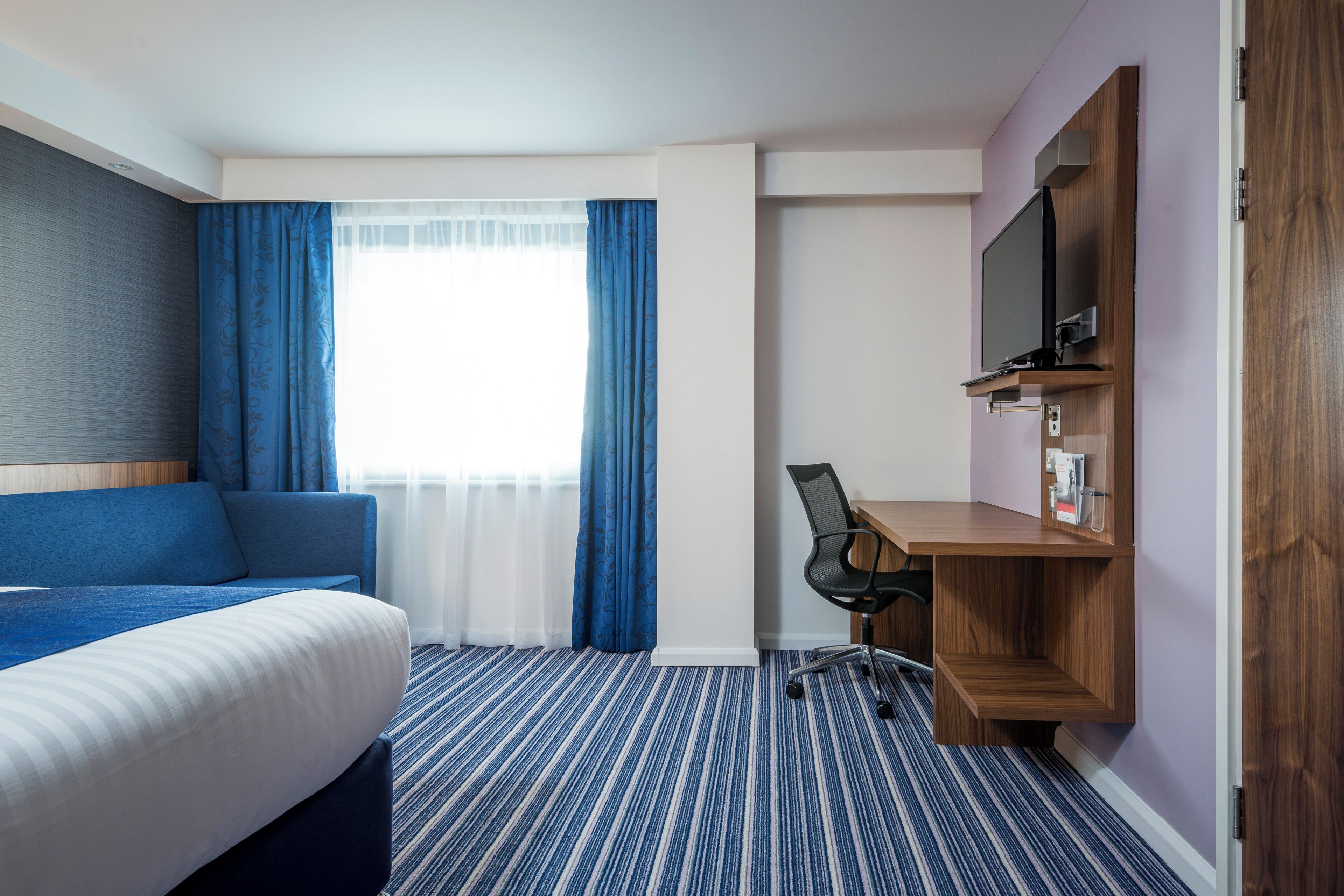 A hotel room with a bed, a desk, a chair, a television, and a window with curtains.