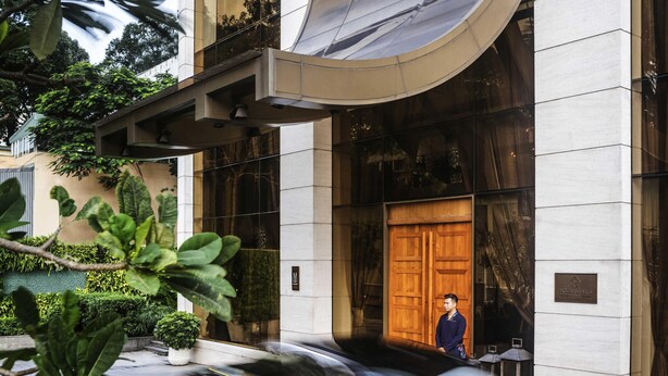 A modern hotel entrance with a large wooden door, a man standing by the door, and a car passing by. - Hôtel des Arts Saigon - MGallery Collection (Ho Chi Minh City)
