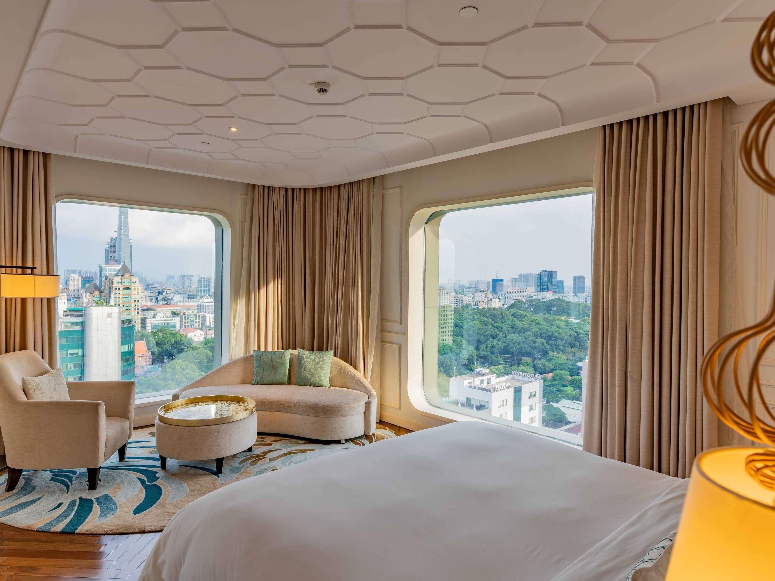 A modern hotel room with a large bed, a curved sofa, a round coffee table, and a view of a cityscape through large windows.