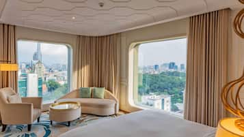 A modern hotel room with a large bed, a curved sofa, a round coffee table, and a view of a cityscape through large windows.