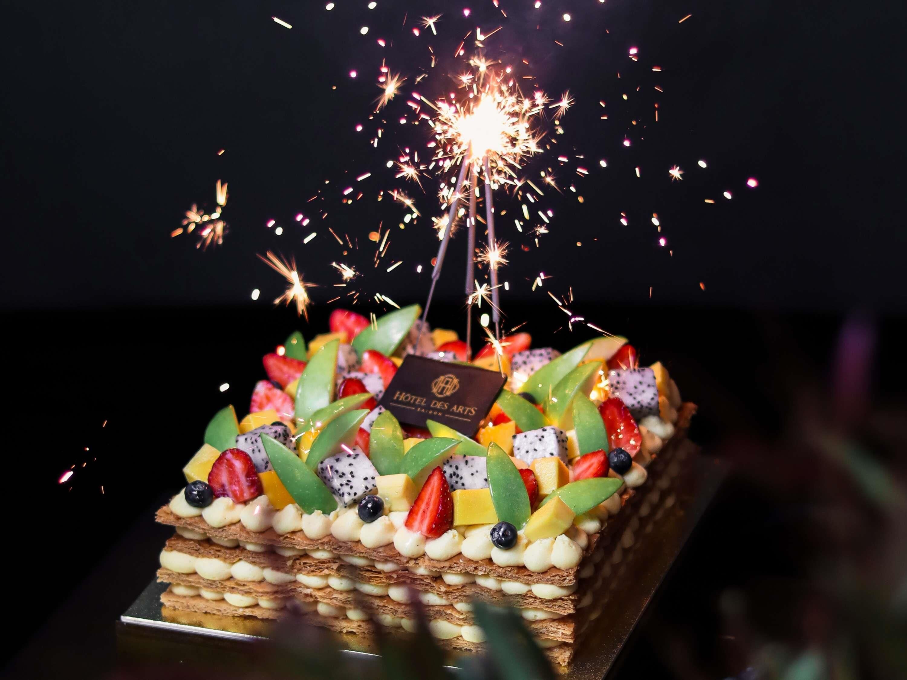 A rectangular cake with multiple layers, topped with various fruits and a sparkler.