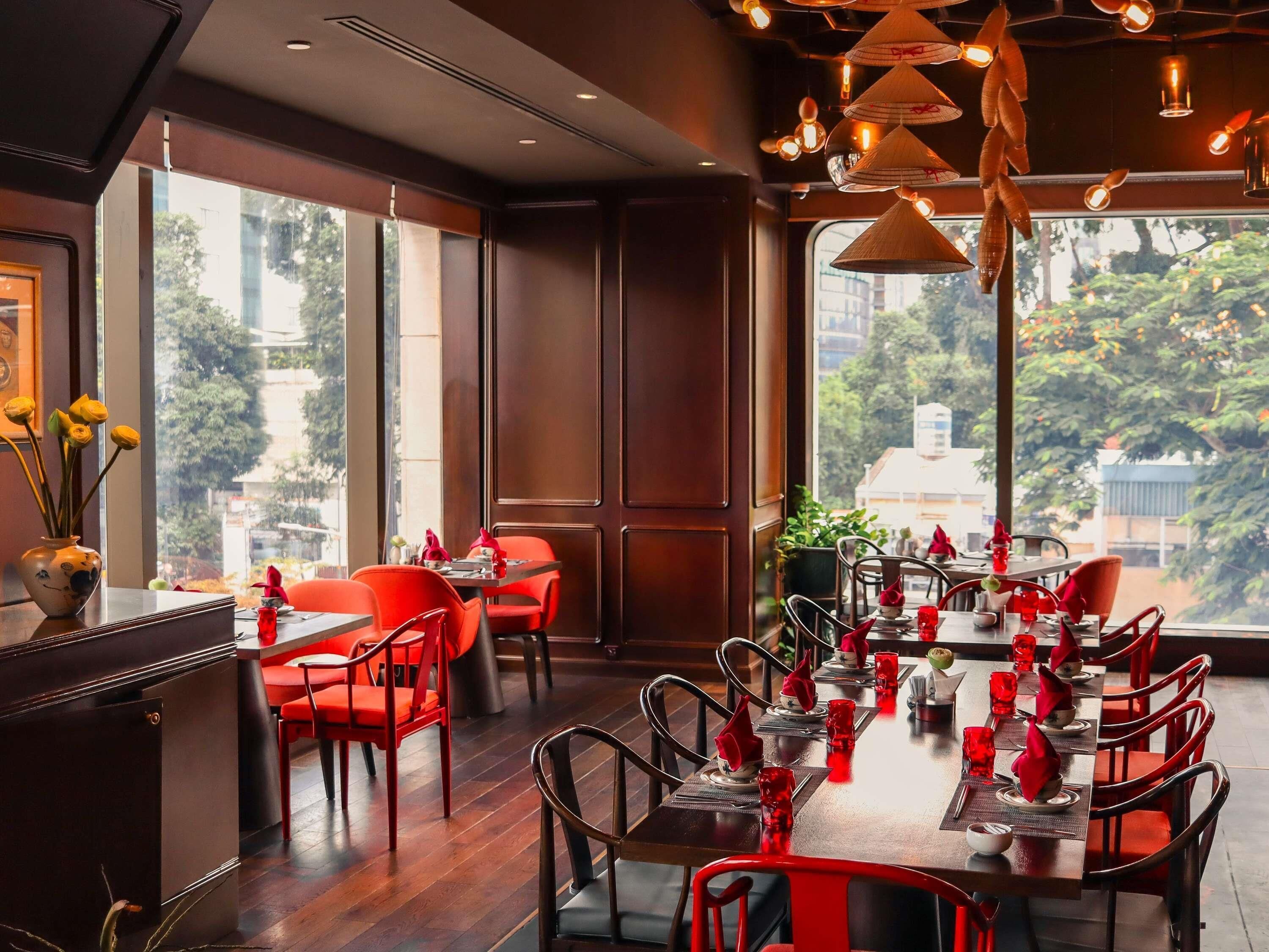 A restaurant with a long dining table, red chairs, and a view of trees through large windows.