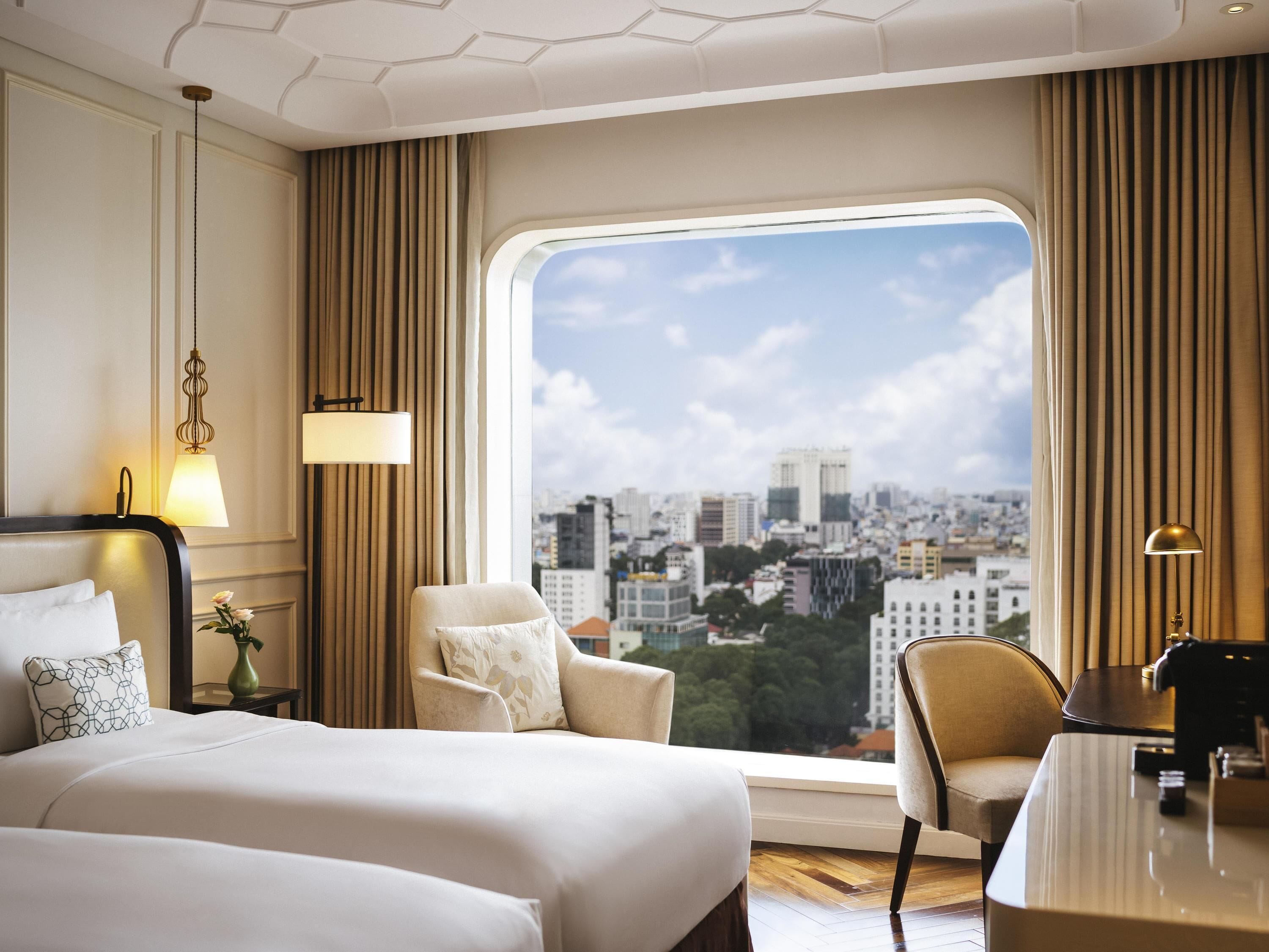 A hotel room with a large window, a bed, a chair, a desk, and a lamp.