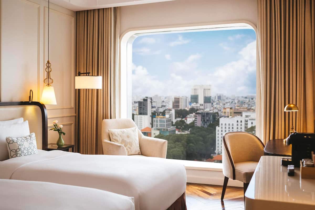 A hotel room with a large window offering a city view, a bed, a chair, a desk, and two lamps.