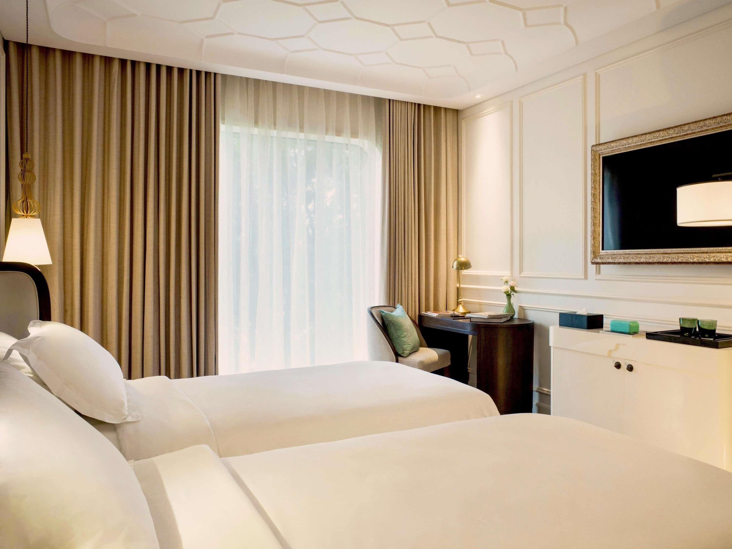 A hotel room with two beds, a desk, a chair, a TV, and a window with curtains.