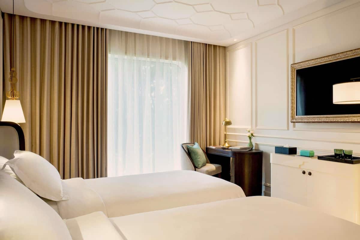A hotel room with two beds, a desk, a chair, a TV, and a window with curtains.