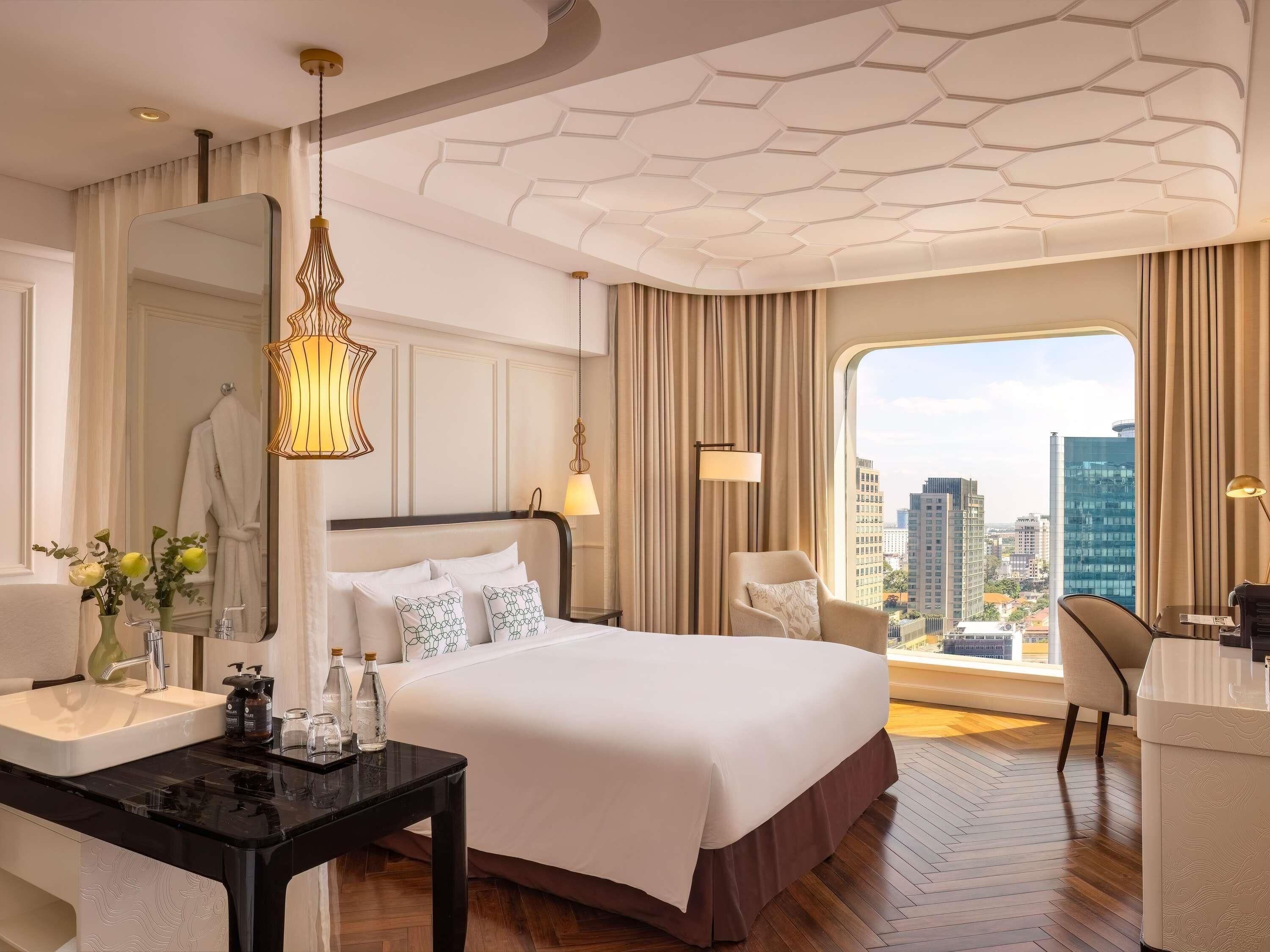 A hotel room with a large bed, a desk, a chair, a view of the city, and a modern ceiling design.