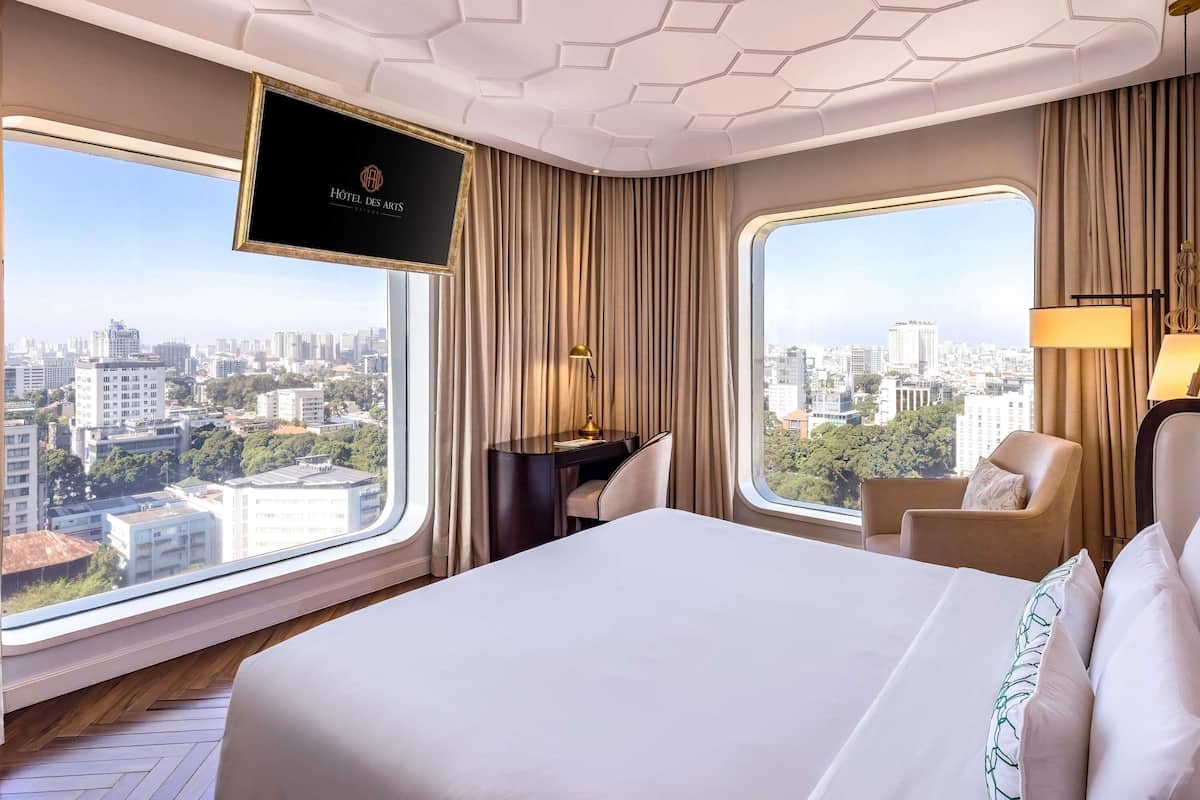 A hotel room with a large bed, a desk with a chair, a TV on the wall, and large windows offering a city view.