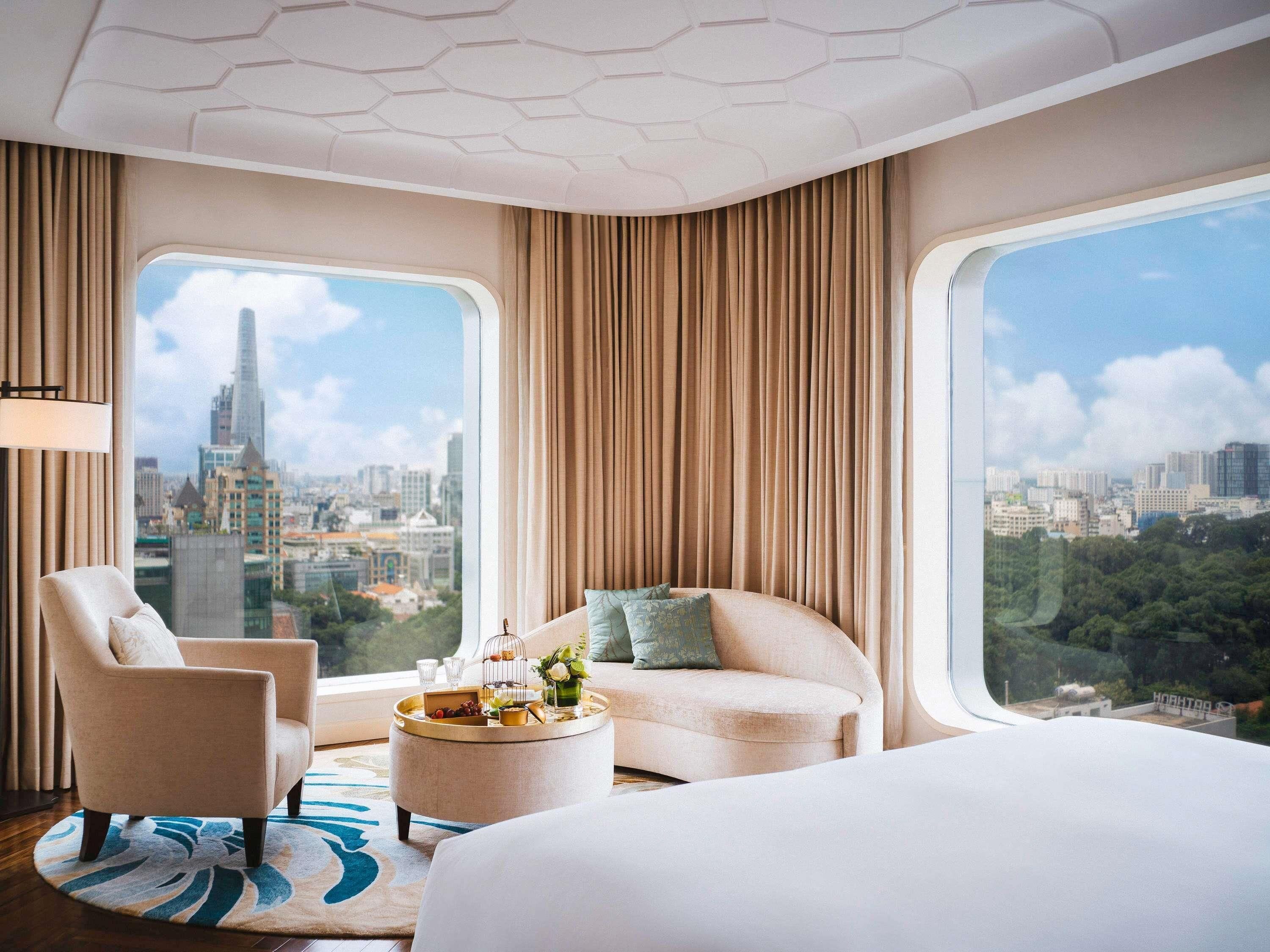 A hotel room with a bed, two armchairs, a sofa, a round coffee table with a tray of snacks, and large windows offering city views.