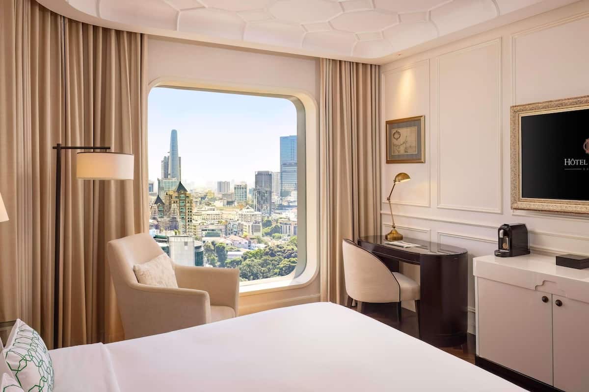 A hotel room with a large bed, a desk, a chair, and a view of a cityscape.