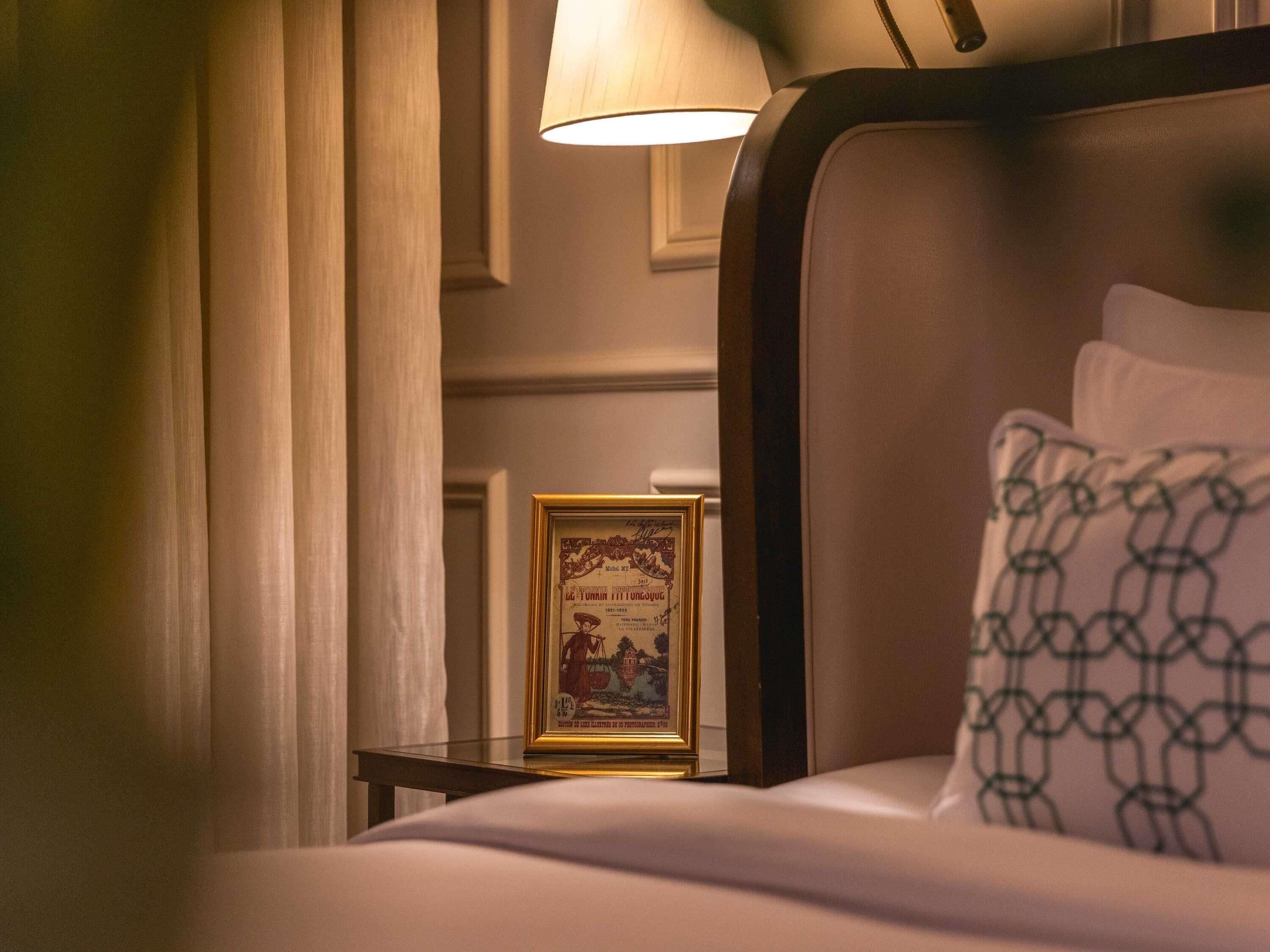 Quarto de hotel com cama, um quadro emoldurado na mesinha de cabeceira e uma luminária.