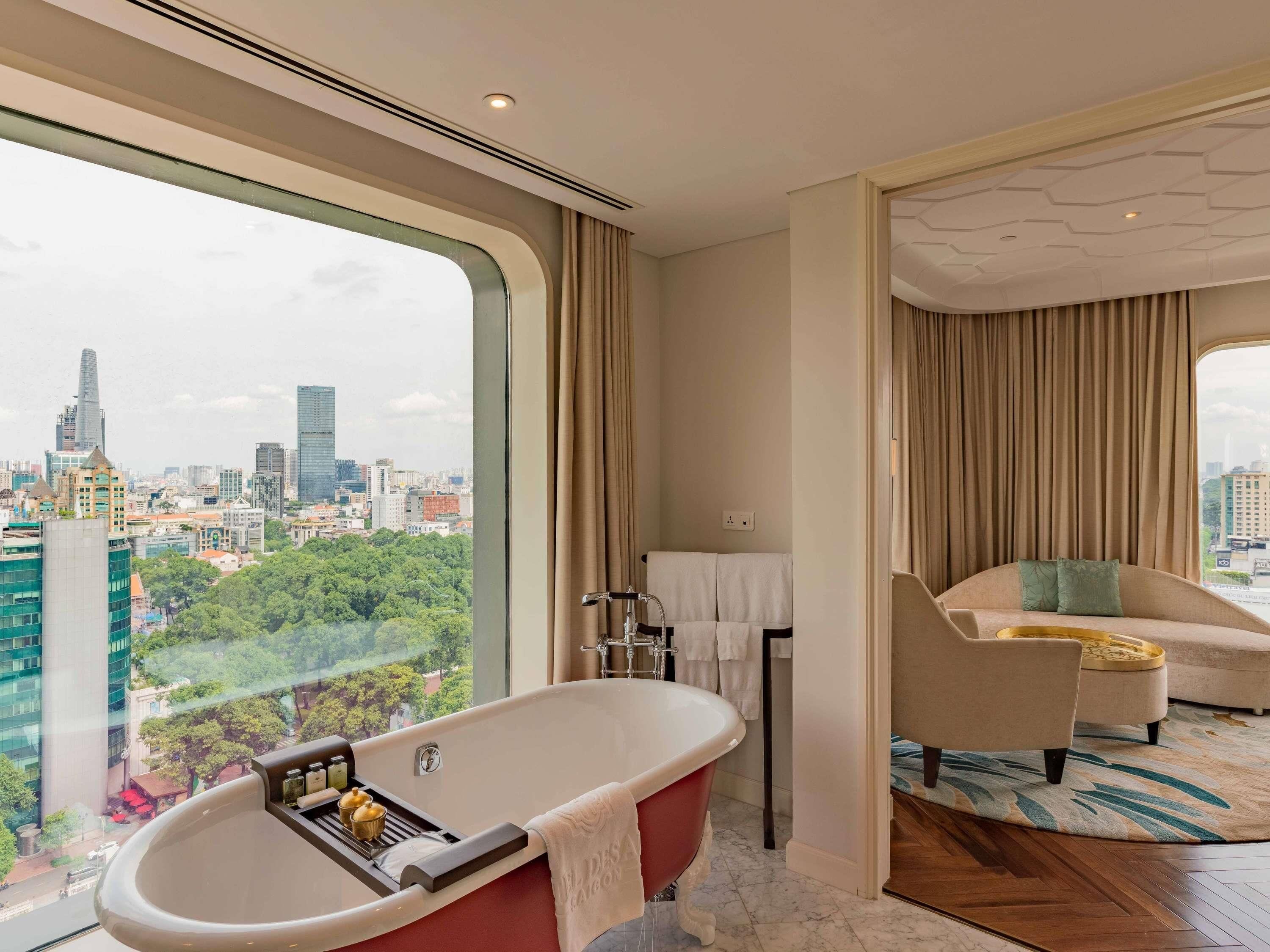 A modern bathroom with a freestanding bathtub, a seating area with a sofa and armchair, and a city view through large windows.